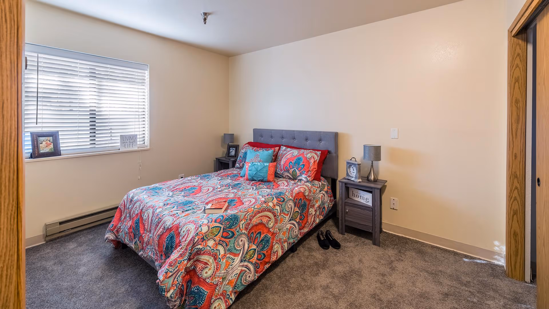 A bedroom with a bed covered in a colorful paisley-patterned comforter and multiple pillows. There are two nightstands on either side of the bed, each with a lamp and framed photos. A window with white blinds is on the left wall, and the floor is carpeted in gray. The walls are painted a light beige color.
