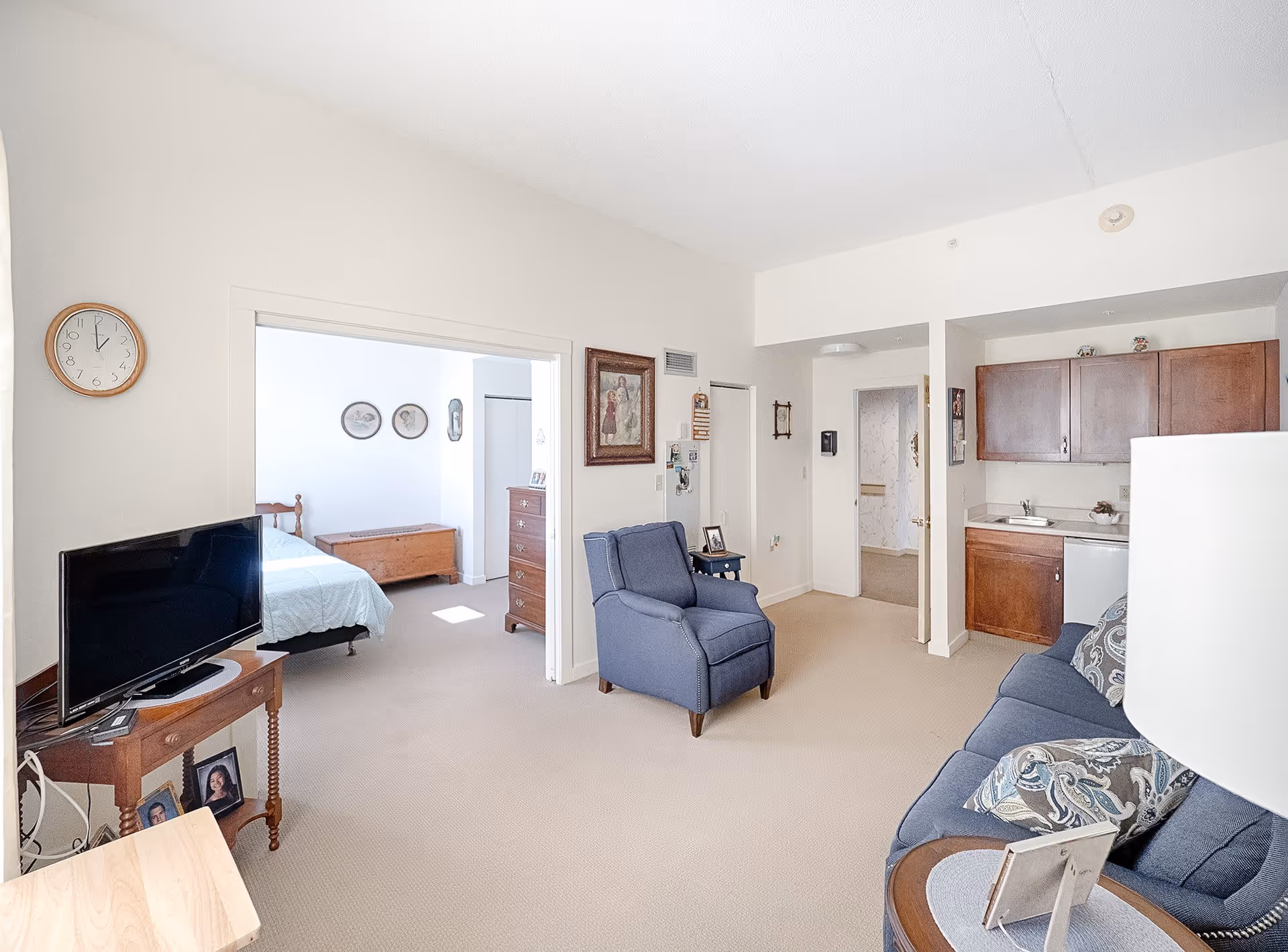A bright and clean senior living apartment featuring a living area with a blue armchair and sofa with patterned pillows, a small wooden table with a TV, and a kitchenette with wooden cabinets and a sink. The bedroom is visible through an open doorway, showing a bed, a wooden chest, and a dresser. The walls are white, and the carpet is beige.