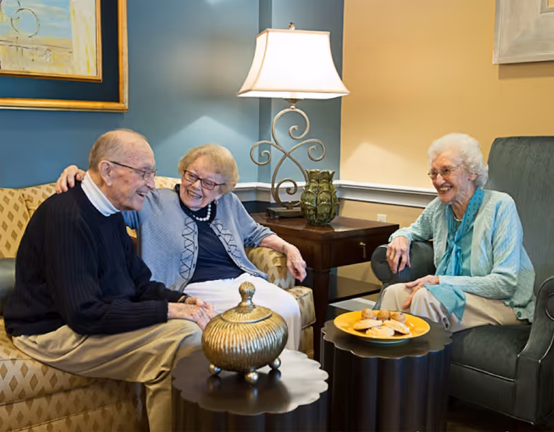 Three elderly people sitting in a cozy living room area, smiling and engaging in conversation. Two women and one man are seated on comfortable chairs and a sofa around two small tables with decorative items and a plate of pastries. A table lamp and framed artwork are visible in the background.