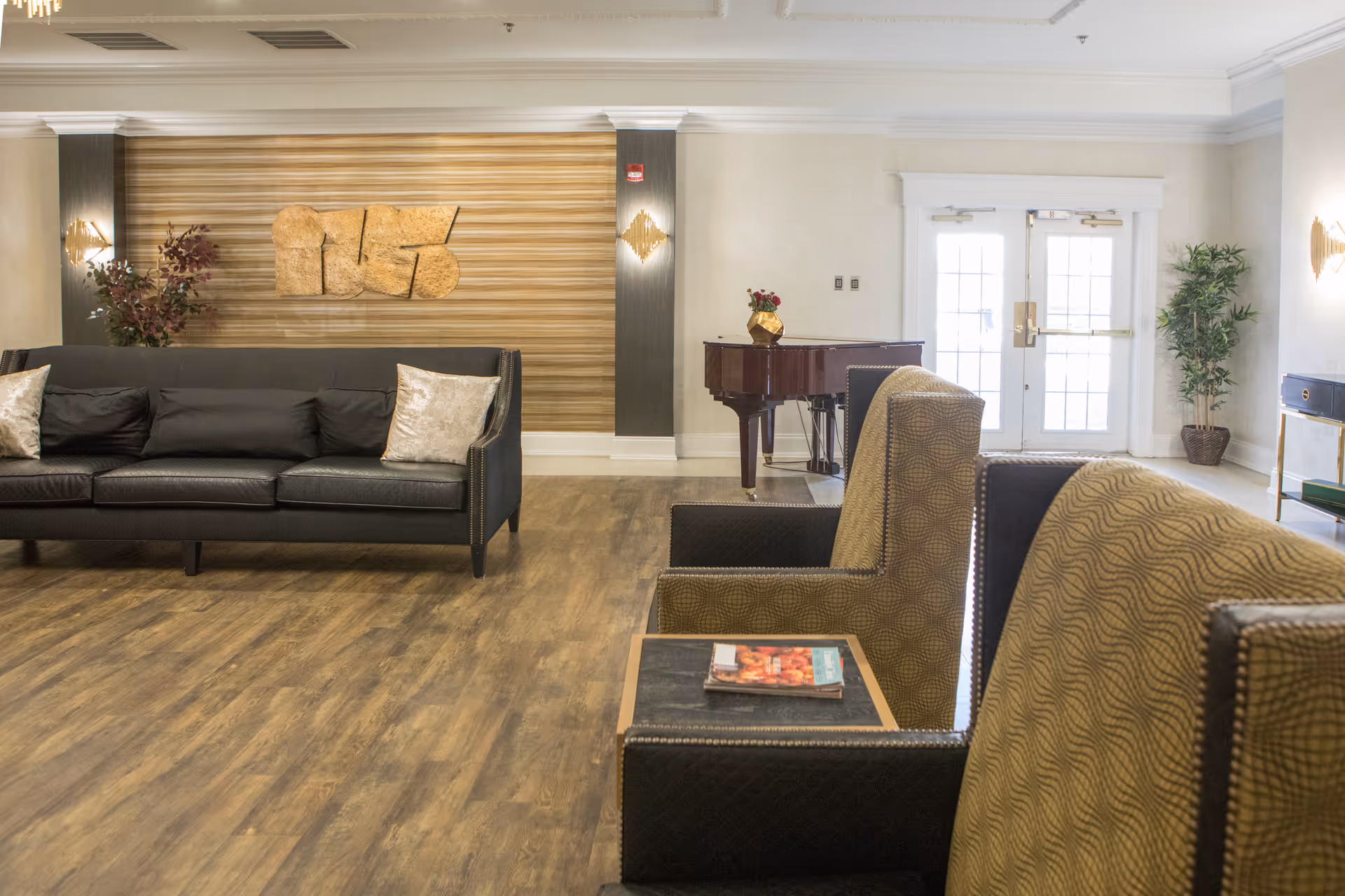 A spacious and well-lit living room area with a black leather sofa adorned with two light-colored pillows, two patterned armchairs with a small table holding magazines between them, a wooden floor, a grand piano in the background, and double glass doors letting in natural light. The walls are decorated with a wooden panel and a large abstract wooden art piece, along with plants and wall sconces.