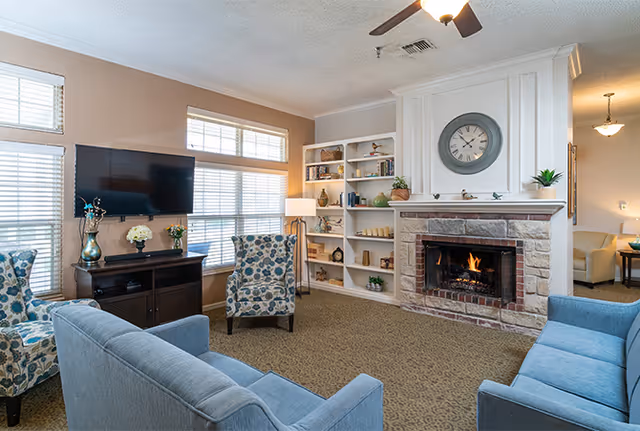 A cozy living room with a lit stone fireplace, a large clock above it, built-in white shelves with decorative items, two blue patterned armchairs, two blue sofas, a flat-screen TV mounted on the wall, and windows letting in natural light.