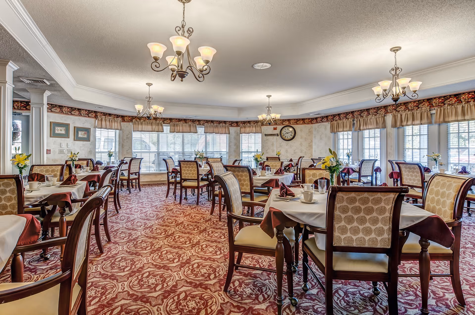 Bright, formal dining room with multiple set tables, upholstered chairs, chandeliers, and patterned carpet.