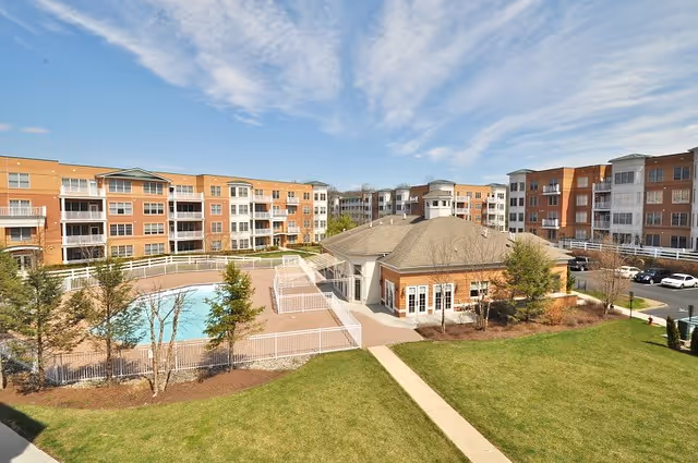 Courtyard view of a senior living complex with a fenced outdoor swimming pool, surrounding brick apartment buildings and a grassy lawn under a blue sky.