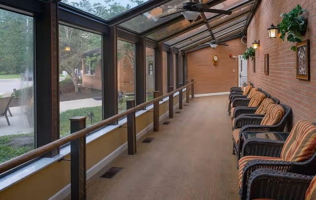 A long enclosed sunroom or hallway with large glass windows and a glass ceiling on one side, overlooking an outdoor area with trees and a patio. The opposite wall is brick with several wall-mounted lights and framed decorations. There are multiple cushioned wicker chairs with striped cushions lined up along the brick wall. Ceiling fans are mounted on the glass ceiling.