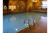 Indoor swimming pool with metal handrails and a brick-tiled floor, surrounded by wooden paneled walls and a small balcony area with railing above one side.