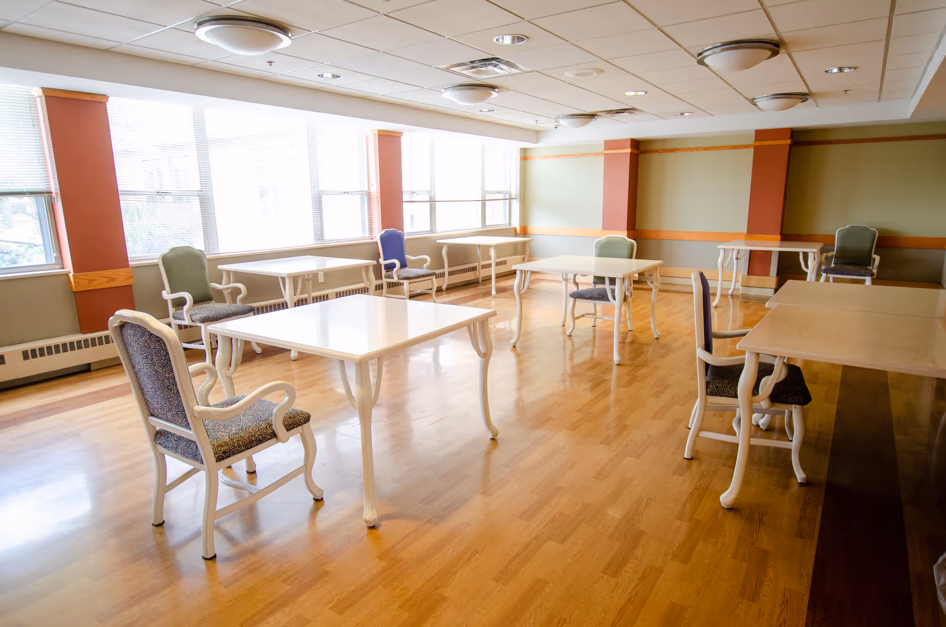 A bright room with several white tables and cushioned chairs spaced apart on a wooden floor, large windows letting in natural light, and a ceiling with recessed lighting fixtures.