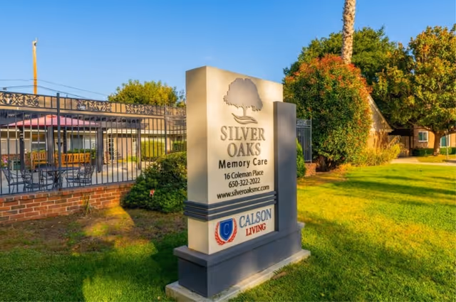 Outdoor view of the Silver Oaks Memory Care facility sign with address, phone number, and website. The sign is placed on a grassy area with trees and a building in the background under a clear blue sky.