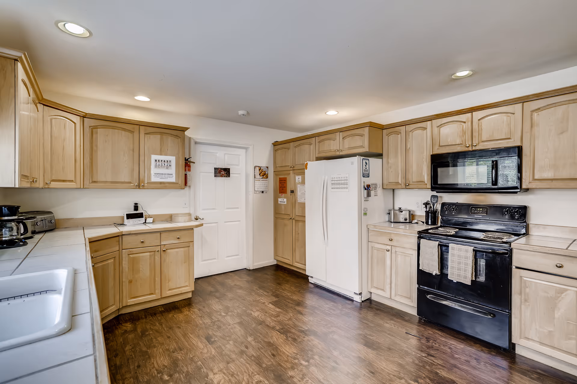 Well-lit kitchen with light wood cabinets, a white refrigerator, black stove and microwave, and dark hardwood floors.