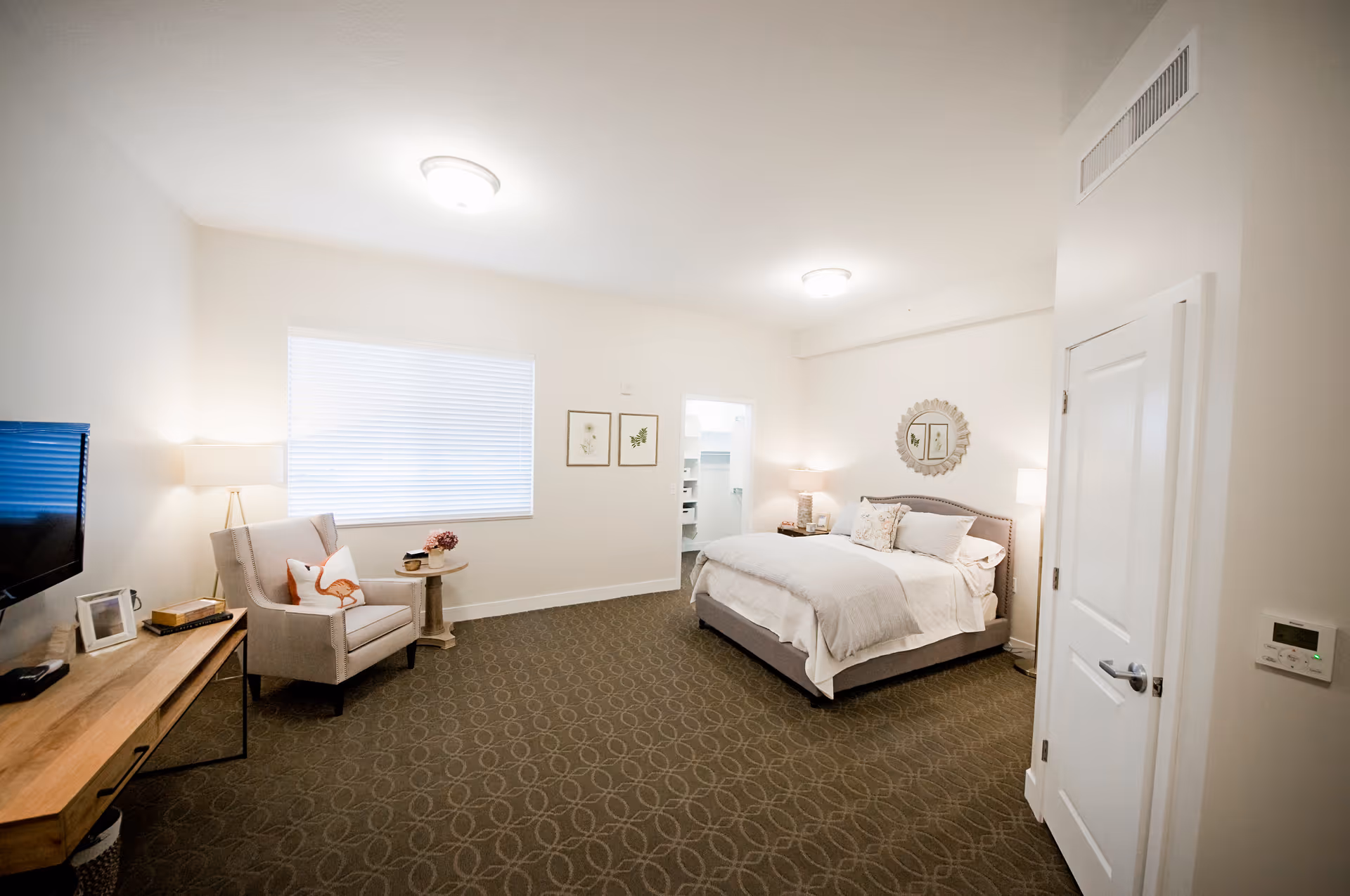 A bright and spacious bedroom in an assisted living facility featuring a large bed with white bedding, a cushioned armchair with a decorative pillow, a wooden console table with a TV, and soft lighting from lamps. The room has beige walls, a patterned carpet, and framed artwork on the walls.