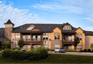 Exterior view of a two-story residential building with stone and beige stucco walls, multiple windows, balconies, and a dark shingled roof under a partly cloudy blue sky. There is a driveway with a parked car and green shrubs and trees in front of the building.