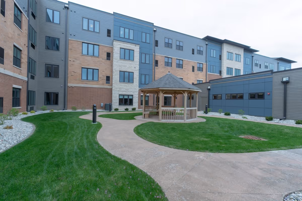 Outdoor courtyard area of a senior living facility with a wooden gazebo in the center, surrounded by green grass and a curved concrete walkway. The multi-story building with a mix of brick and siding is visible in the background under a cloudy sky.