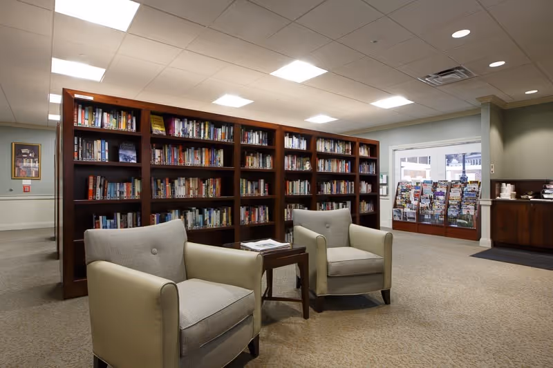 A quiet library area in a senior living facility with two beige armchairs facing a small wooden table. Behind the chairs is a large wooden bookshelf filled with books. To the right, there is a rack with various magazines and brochures near a window, and a coffee station is visible in the corner.