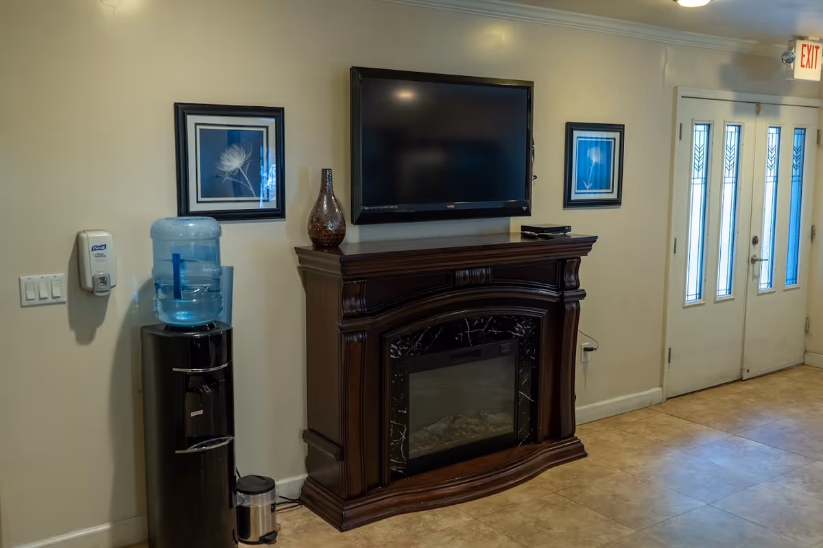 Interior view of a room with a mounted flat-screen TV above a dark wooden electric fireplace. On the left side, there is a black water cooler with a large water bottle on top and a wall-mounted hand sanitizer dispenser. Two framed pictures with floral designs hang on the wall, one on each side of the TV. To the right, there is a double door with frosted glass panels and an exit sign above it. The floor is tiled in a light brown color.