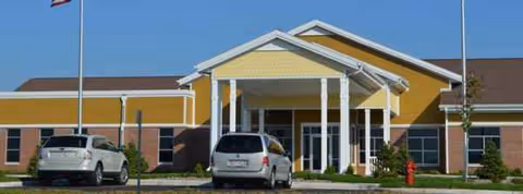 Front view of a yellow senior living facility entrance with a canopy and two parked cars.
