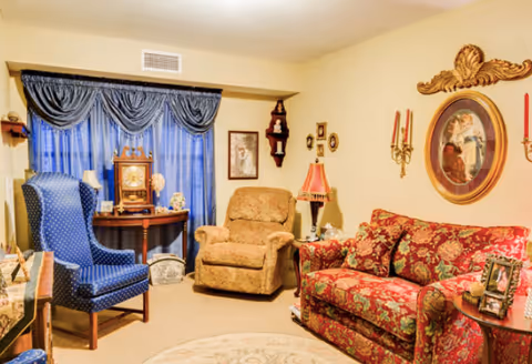 Cozy living room with a patterned red sofa, two upholstered armchairs, a side table and lamp, decorative wall art, and a window with blue drapes.