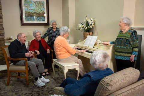 A group of elderly residents gather in a cozy common room as one woman plays a piano and others sit and listen or sing.