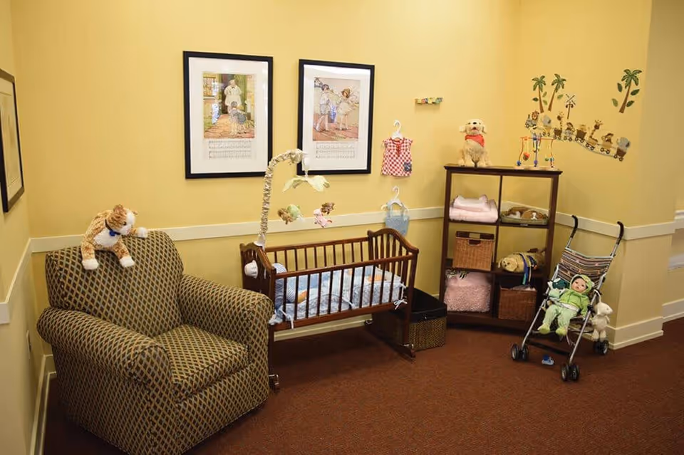 A cozy nursery corner with a patterned armchair, a wooden baby crib with a mobile hanging above it, a small shelving unit with stuffed animals and folded blankets, a doll stroller with a doll dressed in green, and wall decorations including framed pictures and a train-themed wall decal.