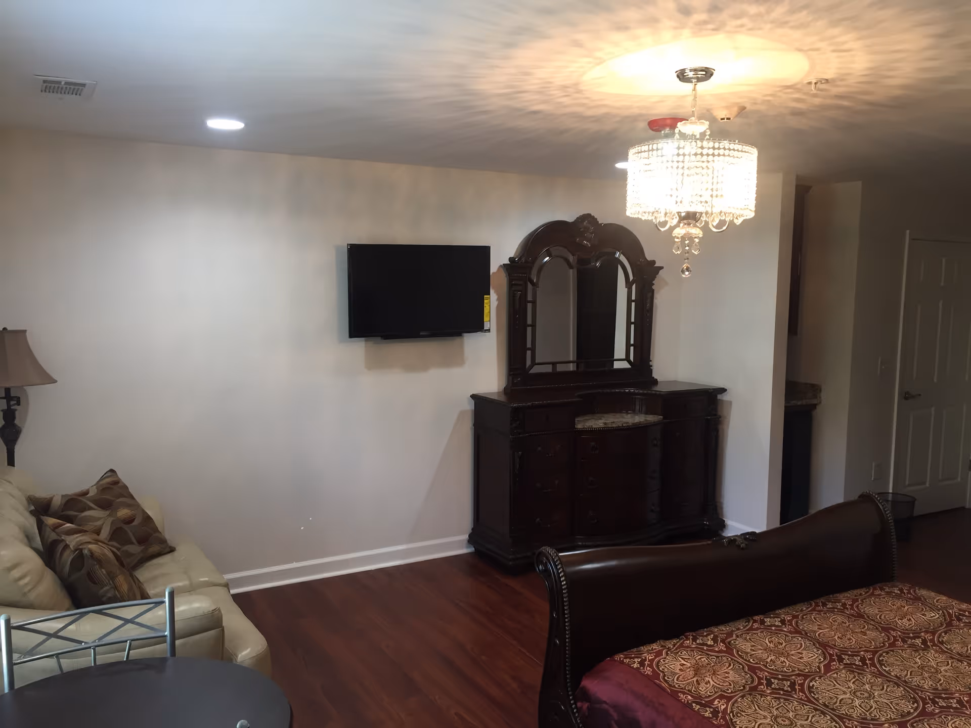 Interior of a room with a wooden floor, a mounted flat-screen TV on a plain wall, a dark wooden dresser with an ornate mirror, a chandelier hanging from the ceiling, a beige leather couch with patterned pillows, and part of a bed with a patterned bedspread.