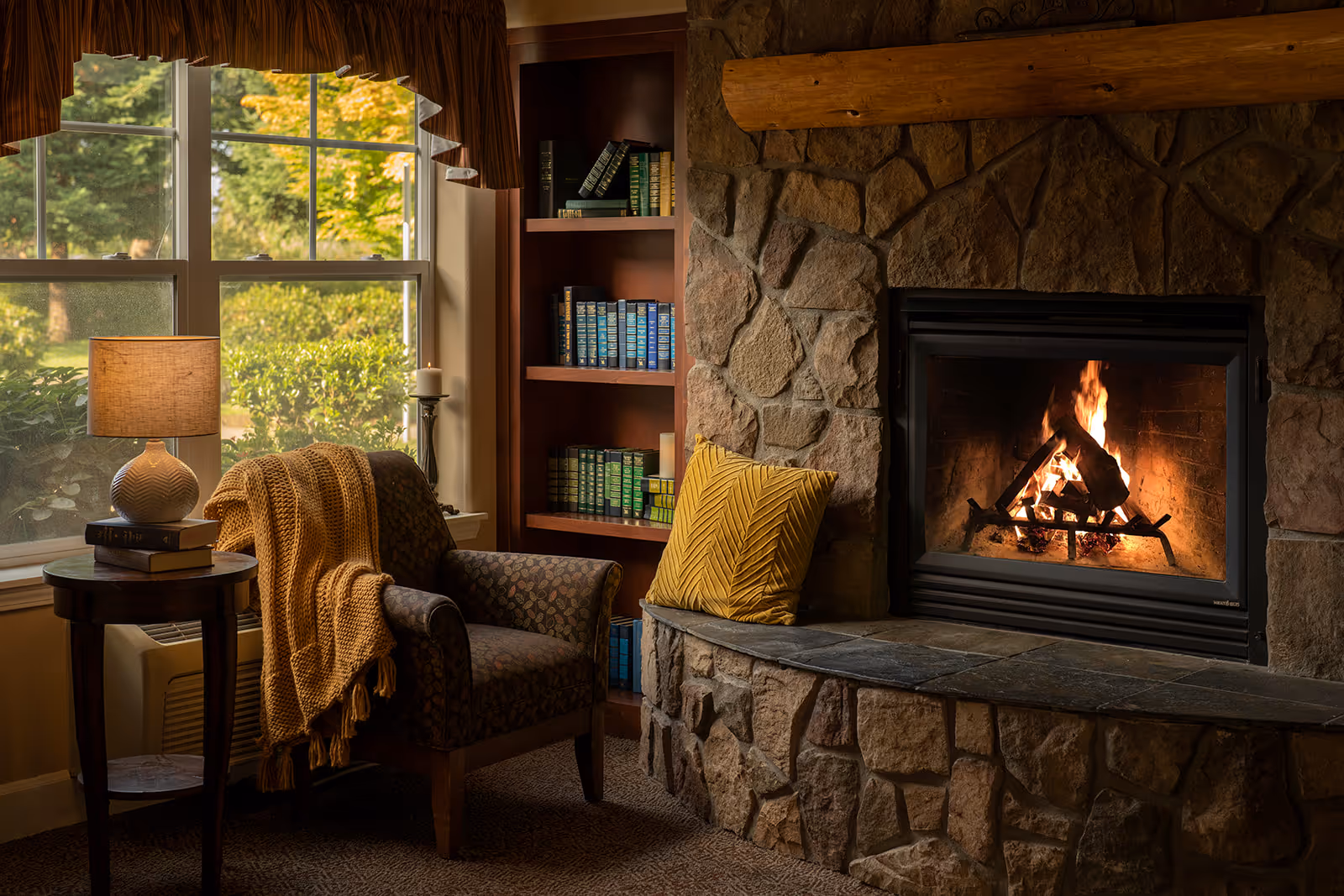 Cozy sitting area with an armchair draped with a blanket, a side table and lamp by a window, and a stone fireplace with a burning fire.