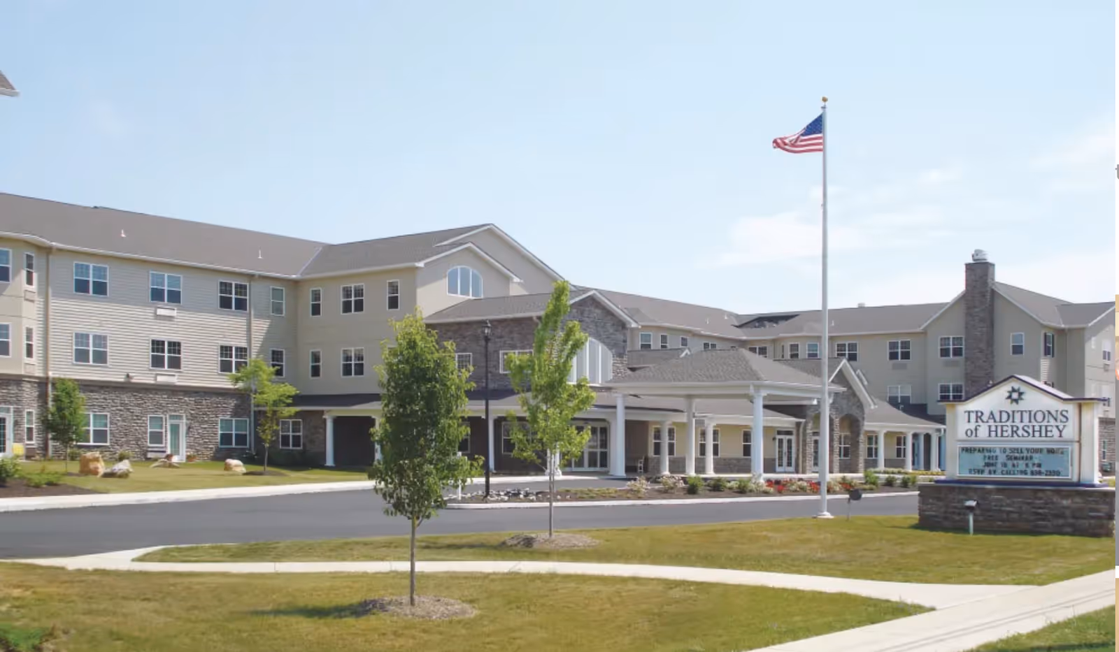 Exterior view of Traditions of Hershey senior living facility showing a large multi-story building with beige siding and stone accents, a covered entrance, an American flag on a tall flagpole, and a sign with the facility name on a grassy lawn with small trees.