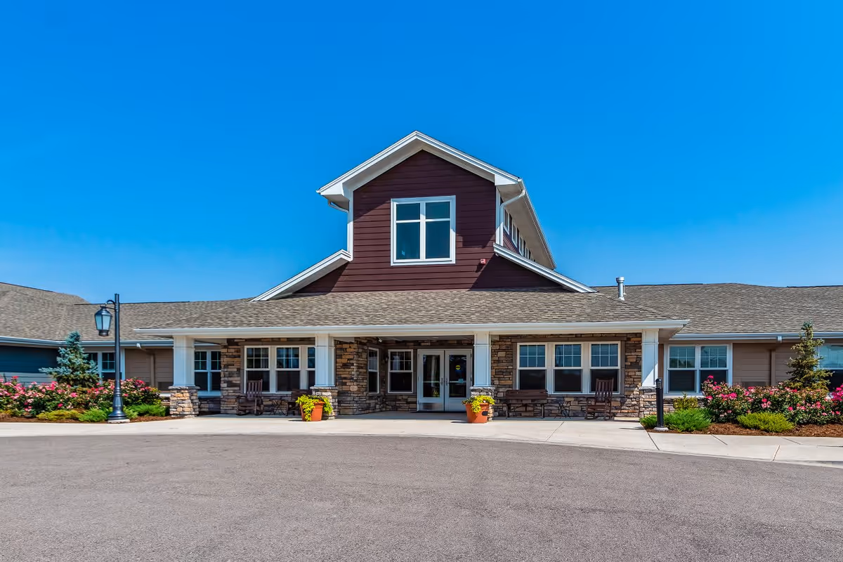 Front exterior view of Willowbrook Place, a single-story building with a gabled roof, stone and wood siding, large windows, and a covered entrance with benches and potted plants on either side. The sky is clear and blue.