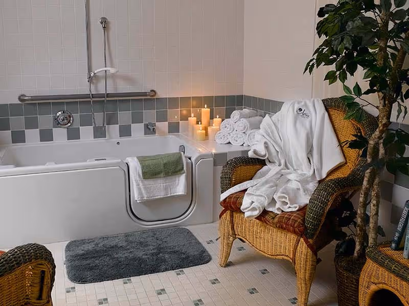 A bathroom featuring a walk-in bathtub with a towel hanging on its door. Several lit candles and rolled white towels are placed on the tub's edge. A wicker chair with a white robe draped over it is positioned next to the tub, and a potted plant is nearby. The floor is tiled, and there is a gray bath mat in front of the tub.