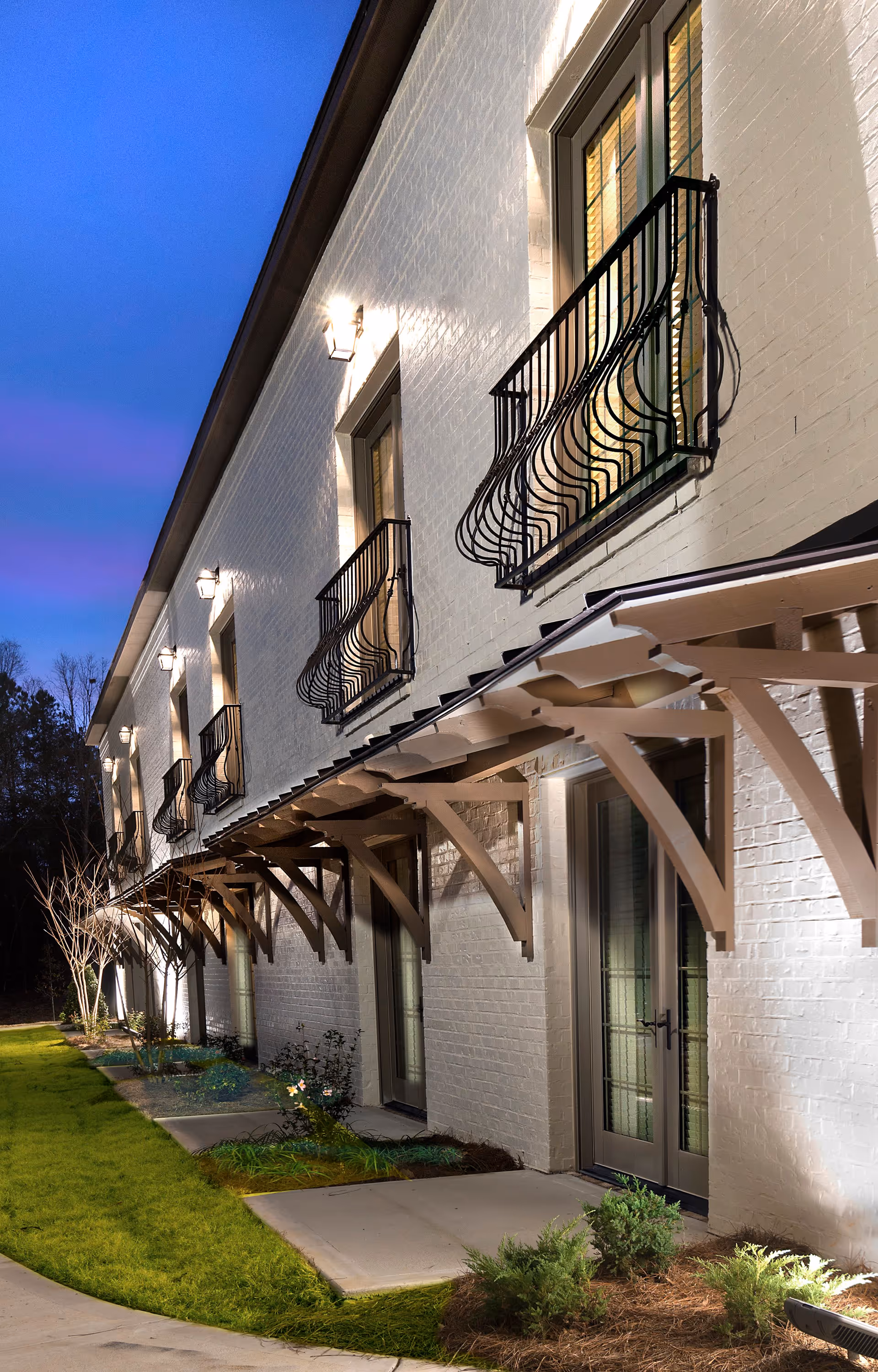 Exterior view of a white brick building at dusk with multiple windows featuring decorative black wrought iron railings and a covered walkway supported by wooden brackets. The building is illuminated by wall-mounted lights and surrounded by a well-maintained lawn and small plants.