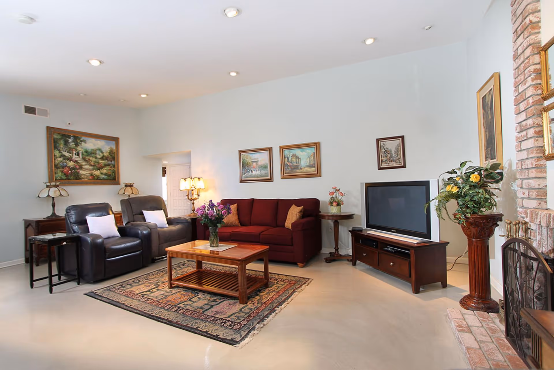 A cozy living room with a burgundy sofa, two black leather armchairs with white pillows, a wooden coffee table with a vase of flowers, a patterned rug, a TV on a wooden stand, several framed paintings on the walls, a brick fireplace, and decorative plants.