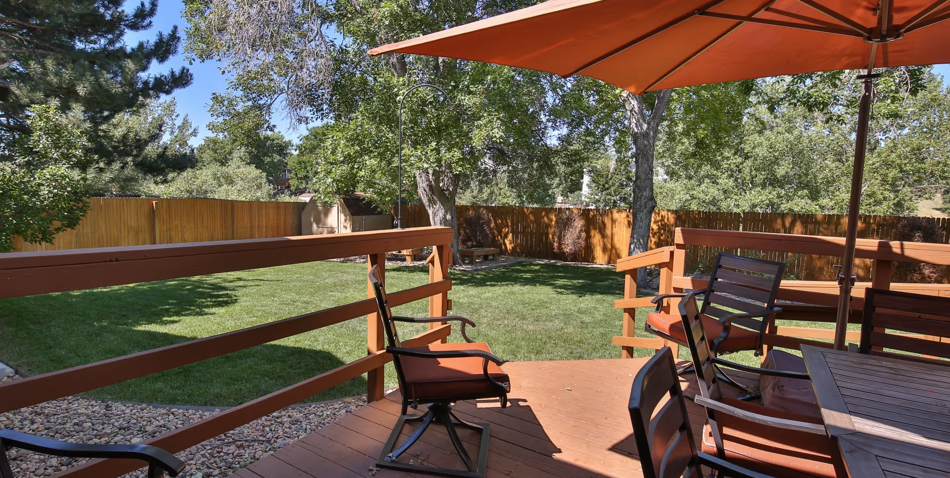 Outdoor patio area with wooden deck, several chairs with orange cushions, a large orange umbrella, and a grassy yard surrounded by a wooden fence and trees in the background.