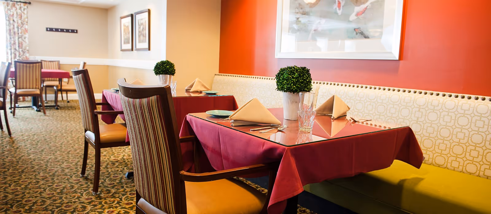A dining area with tables covered in red tablecloths, each set with folded beige napkins, plates, glasses, and silverware. The seating includes striped wooden chairs and a cushioned bench with a patterned backrest against a red and beige wall. A small potted plant decorates each table, and framed artwork hangs on the walls.