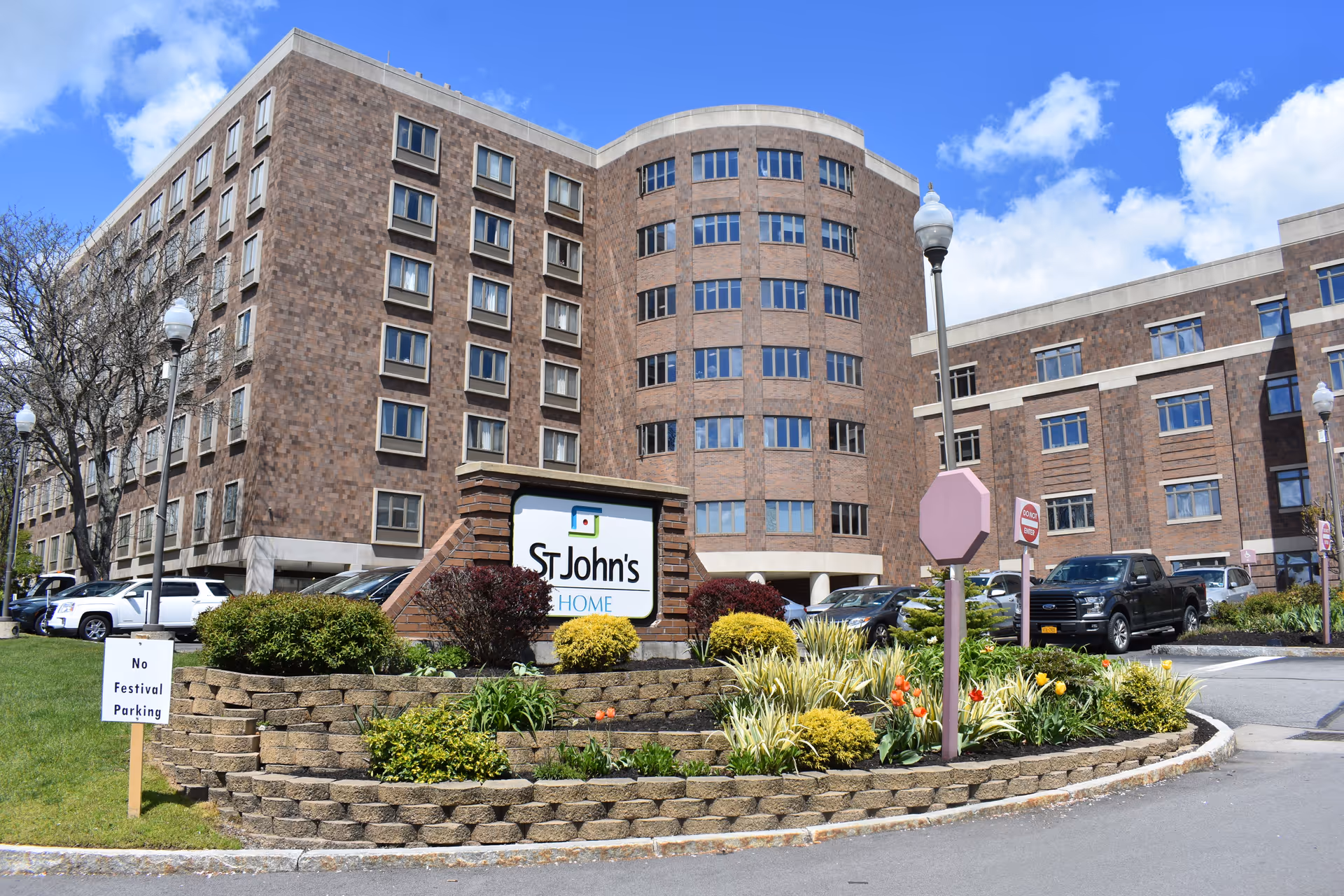 Exterior view of a multi-story brick building with many windows, identified as St. John's Home by a sign in front surrounded by landscaped bushes and flowers. Several cars are parked in the parking lot around the building under a partly cloudy blue sky.