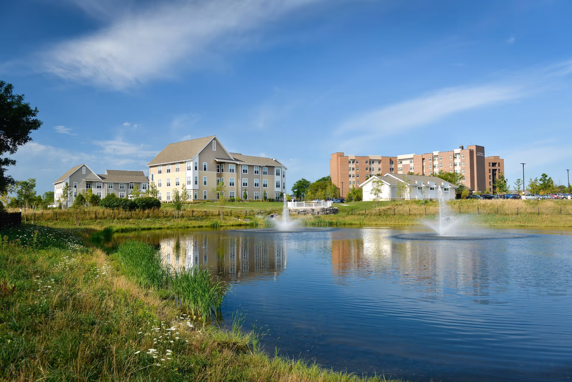 A scenic view of the Jewish Home facility showing multiple buildings near a pond with two water fountains. The sky is clear with some clouds, and there is greenery surrounding the pond and buildings.