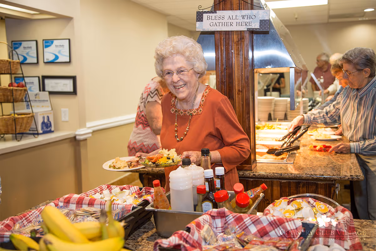 An elderly woman smiles while serving food from a buffet in a communal dining area.