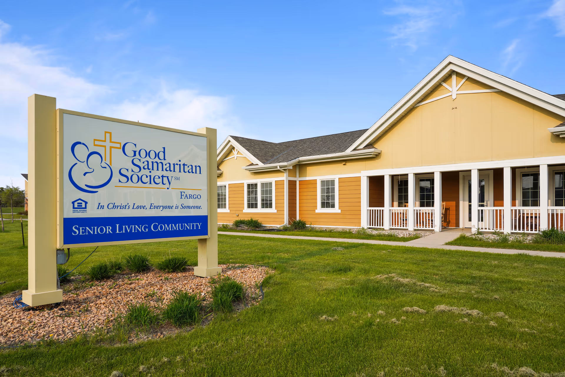 Exterior view of Good Samaritan Society - Fargo senior living community building with a large sign in the foreground displaying the facility's name, logo, and tagline. The building is yellow with white trim and has a covered porch with railings. The surrounding area has green grass and a clear blue sky.