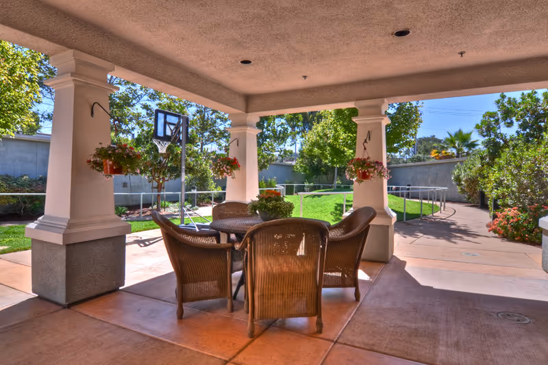 Covered outdoor patio area with a round table and four wicker chairs surrounded by hanging flower pots. The patio overlooks a green lawn with trees, a basketball hoop, and a paved walkway with a handrail.