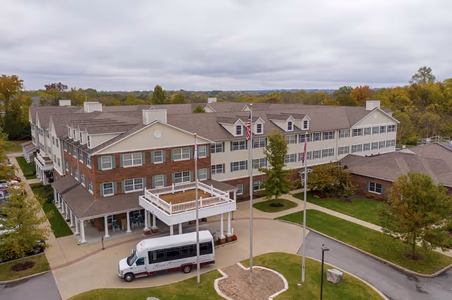 A large three-story senior living facility building with a combination of brick and beige siding exterior. The building has multiple windows and a covered entrance with a balcony above it. There are three flagpoles with flags in front of the building and a white shuttle bus parked near the entrance. Surrounding the building are trees and landscaped green areas under a cloudy sky.