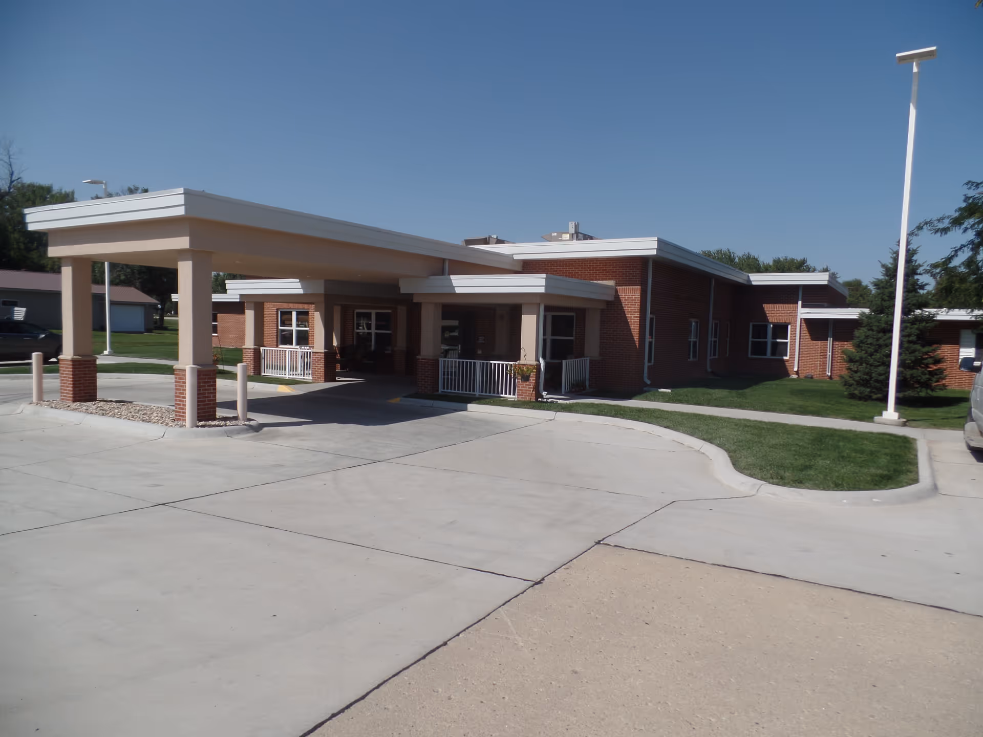 Exterior view of Pleasant Acres Care Center showing a single-story brick building with a covered entrance driveway, surrounded by green grass and a clear blue sky.