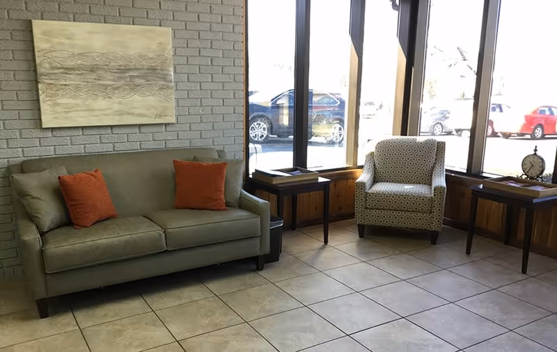 A seating area with a green sofa adorned with two orange cushions, a patterned armchair, two wooden side tables, and large windows showing parked cars outside. A decorative painting hangs on the light gray brick wall behind the sofa.