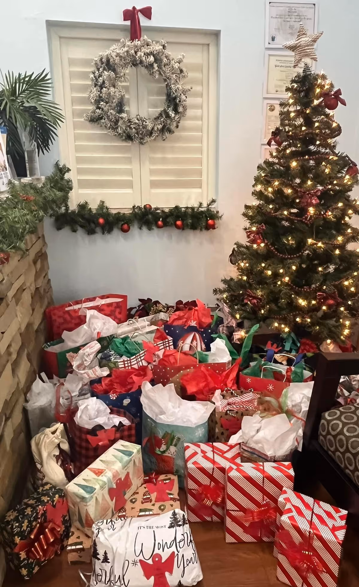 A festive indoor scene featuring a decorated Christmas tree with lights and a star topper, surrounded by numerous wrapped gift bags and boxes. A frosted wreath with a red bow hangs on a white shuttered window above a garland decorated with red ornaments. Part of a stone wall and a patterned chair are visible on the sides.