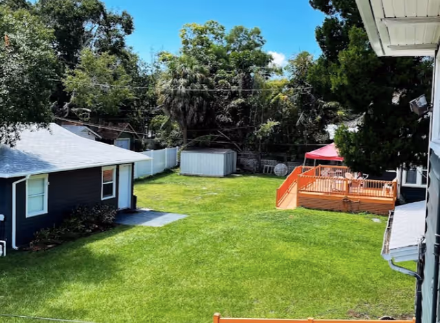 View of a spacious backyard with green grass, a small shed, a wooden deck with a red canopy, and surrounding trees under a clear blue sky.