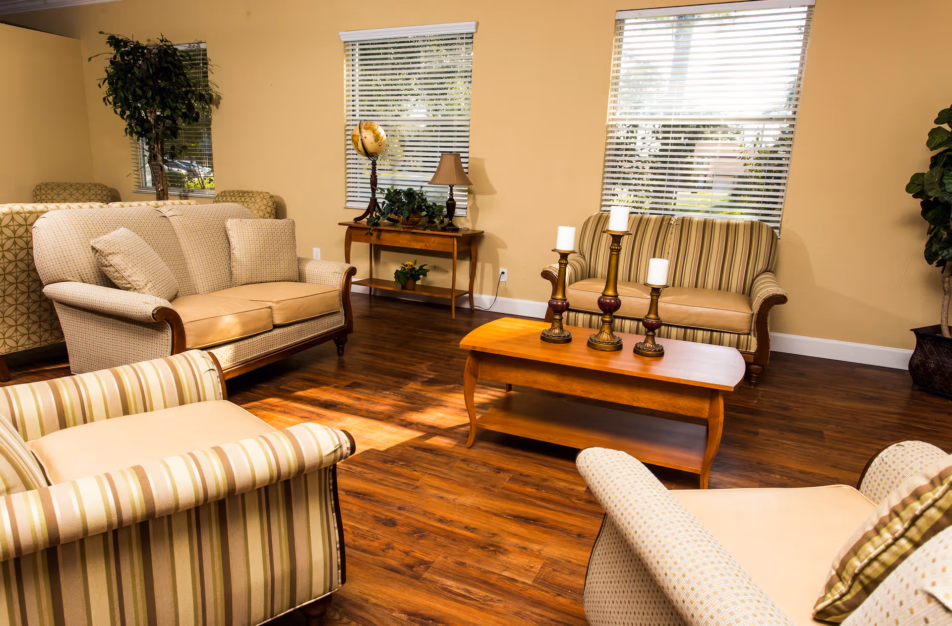 A cozy living room with wooden flooring, featuring two beige patterned sofas, two striped armchairs, a wooden coffee table with three candle holders, a wooden side table with a globe, lamp, and plants, and two windows with white blinds letting in natural light.