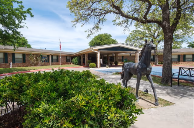 Exterior view of a single-story brick building with a covered entrance, surrounded by green bushes and trees. A black horse statue is prominently displayed near the entrance on a paved walkway. A bench is visible to the right under a tree, and an American flag is flying near the building.