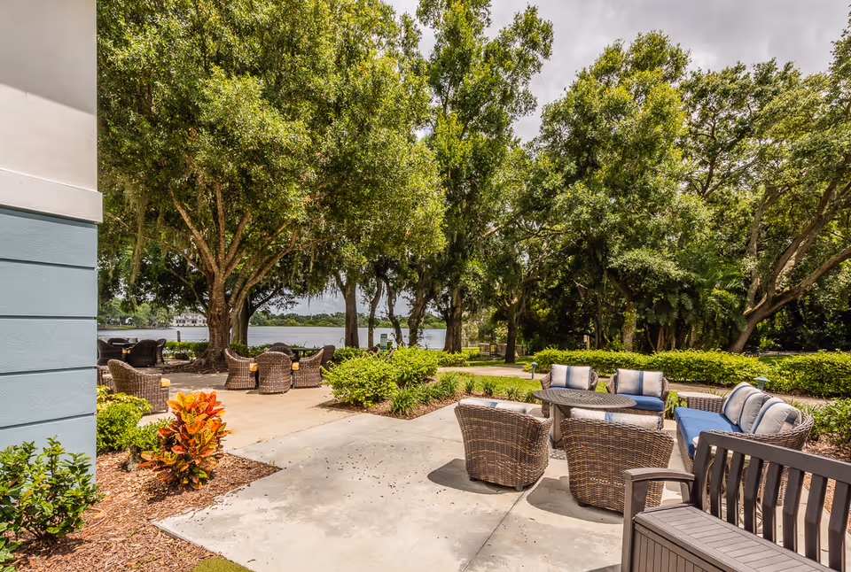 Outdoor patio area with wicker chairs and cushioned sofas arranged around small tables, surrounded by lush green trees and bushes, with a view of a lake in the background under a partly cloudy sky.