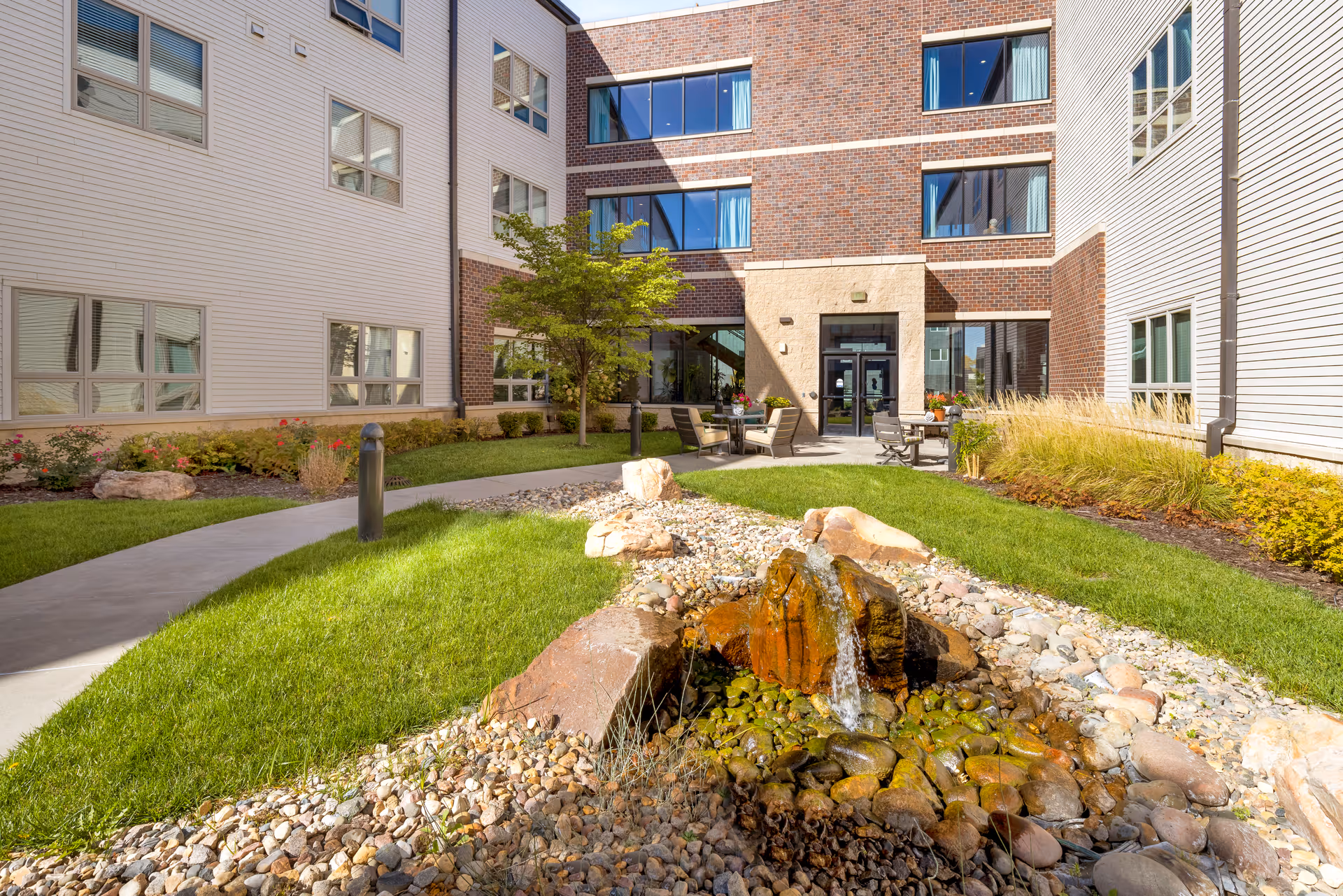 Outdoor courtyard area of a senior living facility with a small rock water feature in the foreground, green grass, a paved walkway, and patio seating with tables and chairs. The building surrounding the courtyard has multiple windows and a mix of brick and siding exterior.