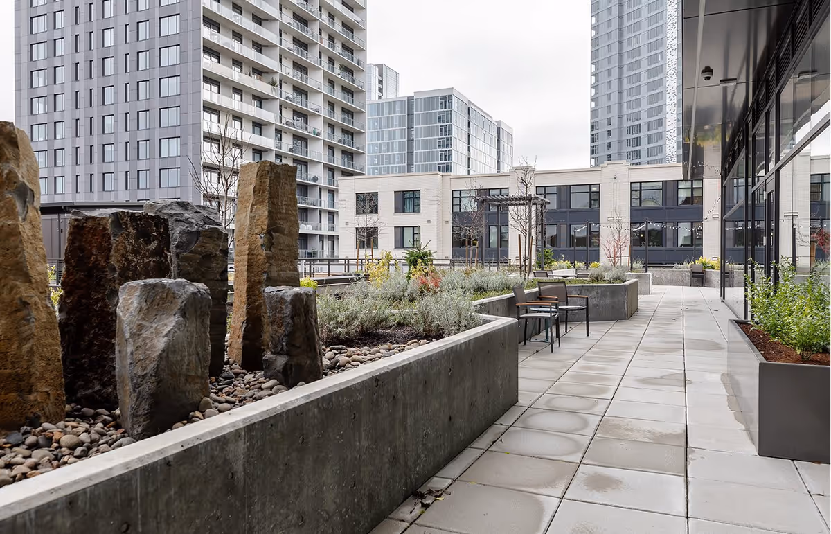 Outdoor patio area with concrete planters containing rocks and plants, several chairs, and modern high-rise buildings in the background under an overcast sky.