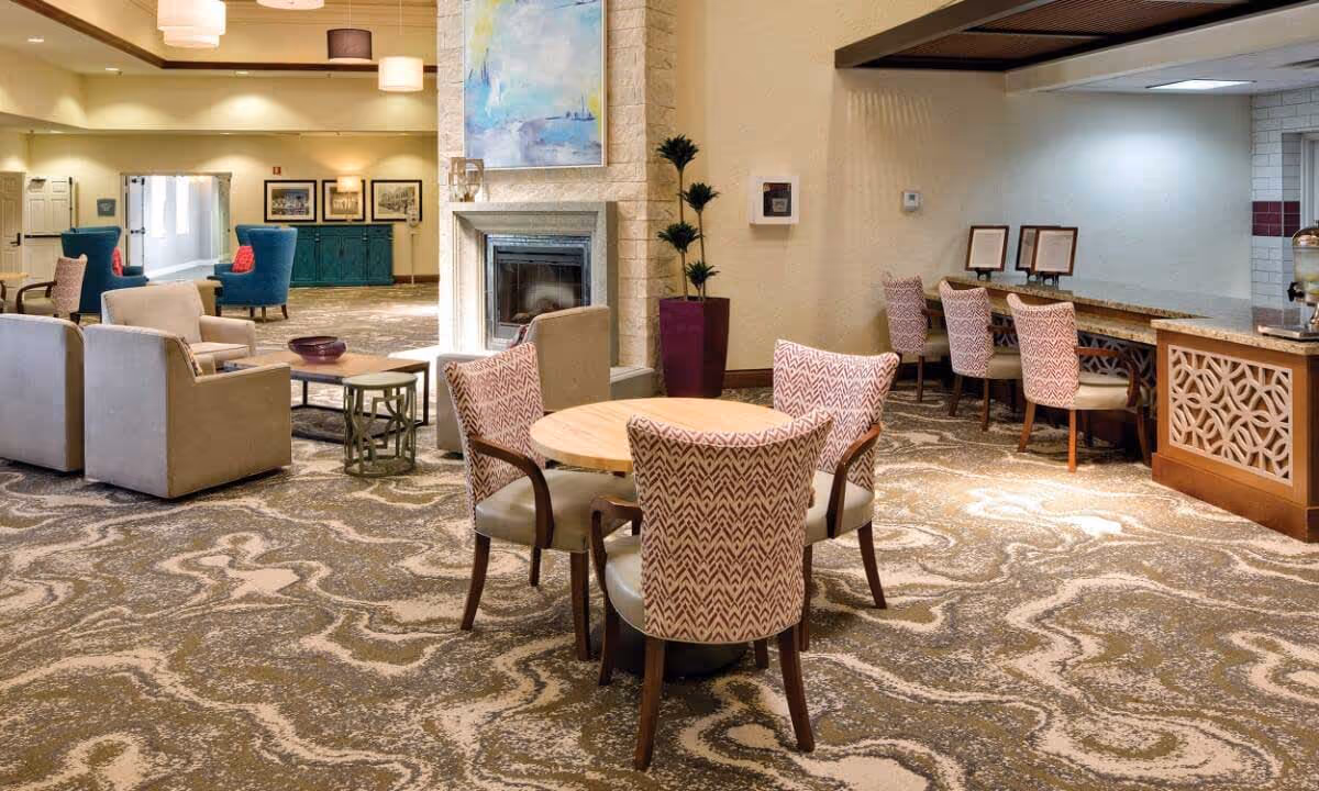 A spacious and well-lit common area in a senior living facility featuring a round wooden table with four patterned chairs in the foreground. In the background, there are several armchairs arranged around a coffee table near a fireplace with a large abstract painting above it. To the right, a counter with three high chairs is visible, along with a decorative wooden panel and a water dispenser. The room has patterned carpeting and soft lighting.