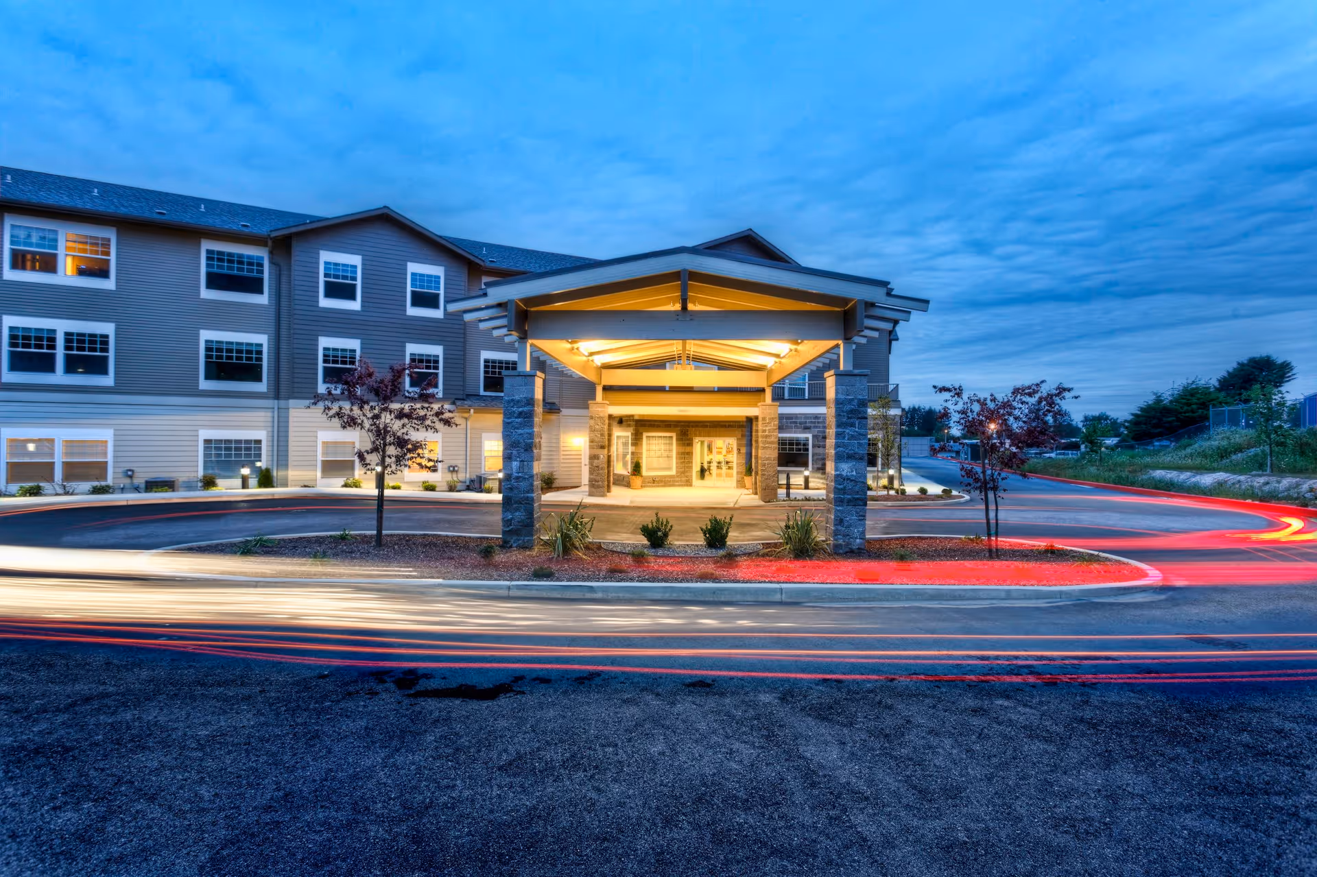Exterior view of a senior living community building at dusk with a covered entrance illuminated by warm lights. The building has multiple windows and a driveway with light trails from passing vehicles.