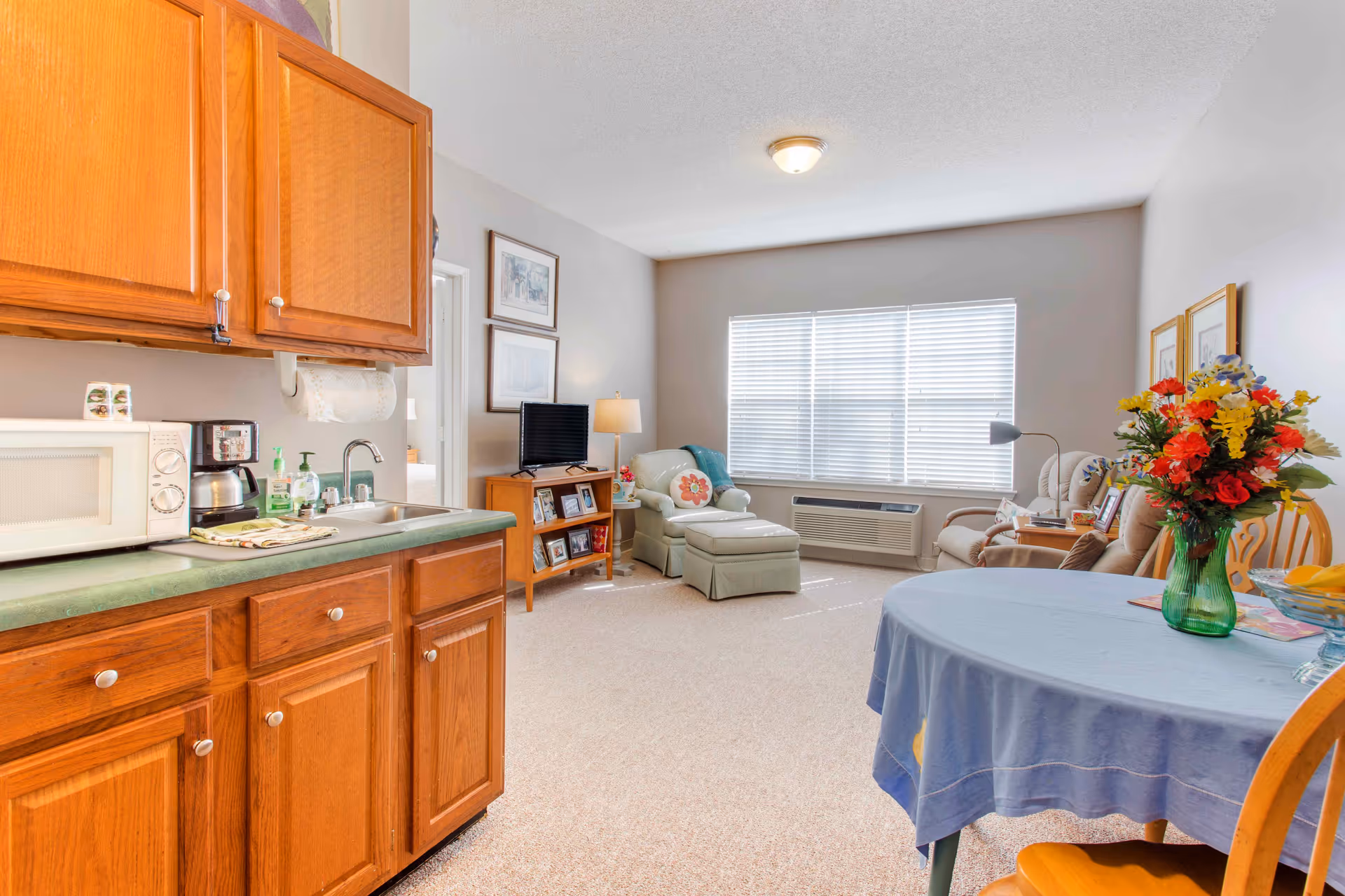 Open studio-style living area with a small kitchenette at left, dining table with flowers, seating area, TV stand and a large window.