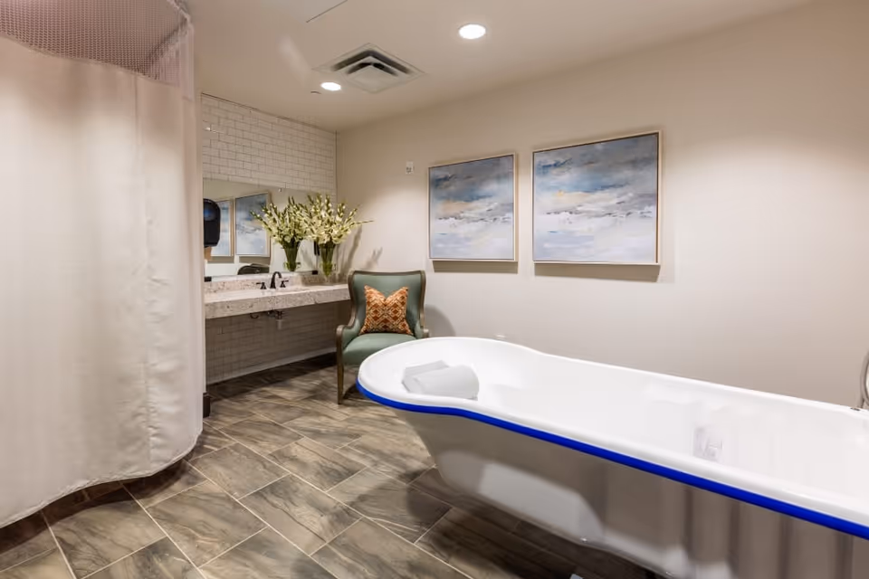 A spacious bathroom featuring a white bathtub with a blue rim, a green armchair with an orange patterned pillow, a vanity with a granite countertop and sink, a large mirror, a vase with white flowers, two abstract paintings on the wall, and a shower area with a beige curtain.