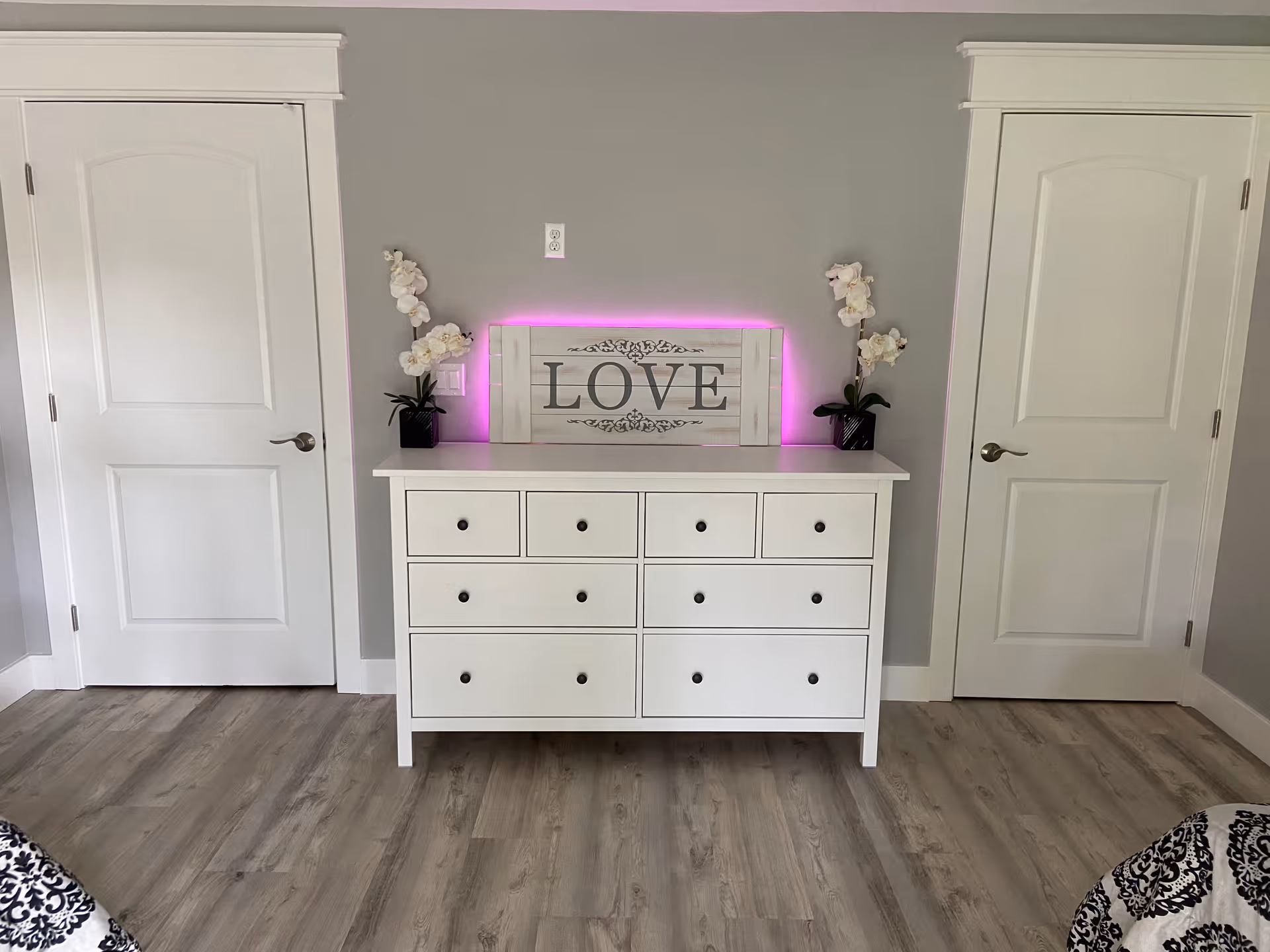 A white dresser with multiple drawers stands against a gray wall between two white doors. On top of the dresser is a decorative sign with the word 'LOVE' illuminated by a soft pink backlight, flanked by two white orchid plants in black pots. The floor is a light wood laminate, and parts of a black and white patterned bedspread are visible at the bottom corners of the image.